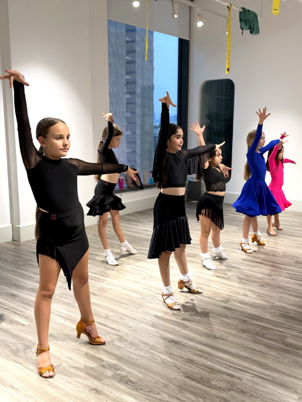 Girls in Latin dance outfits practising arm positions in a bright dance studio